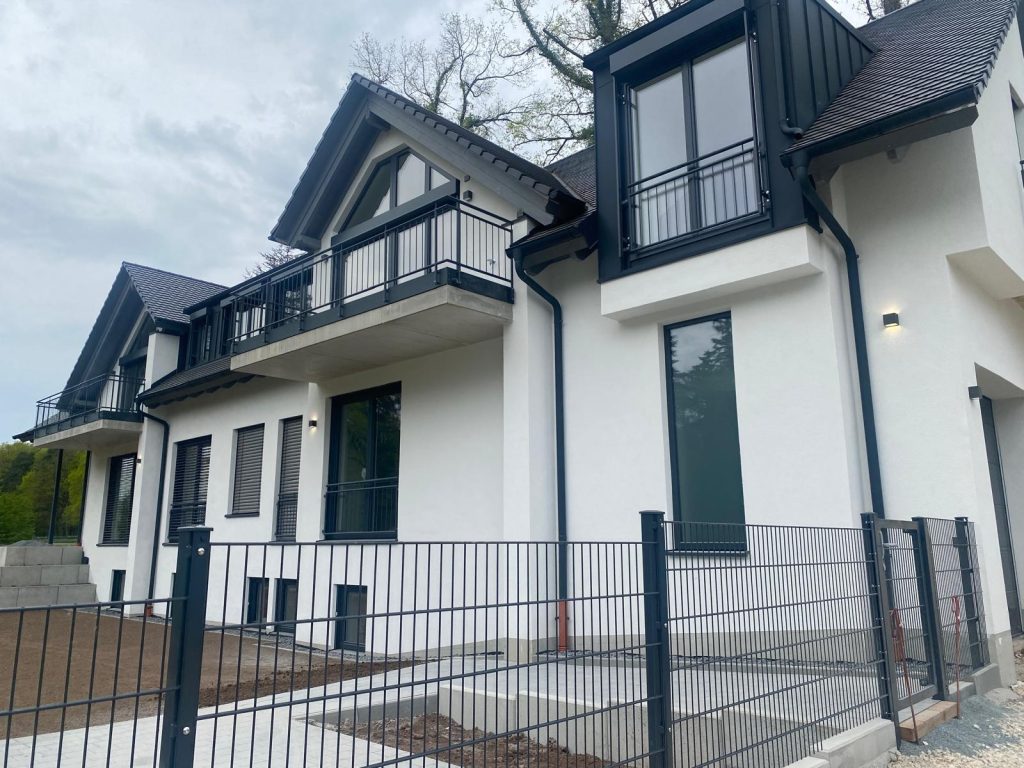 Modernes weißes Haus mit schwarzen Akzenten, Balkonen, großen Fenstern und einem umzäunten Garten vor bewölktem Himmel.