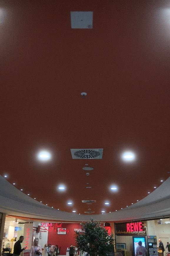 Blick in das Innere eines modernen Einkaufszentrums mit roter Decke, kreisförmiger Beleuchtung, Belüftung und sichtbaren Ladeneingängen.